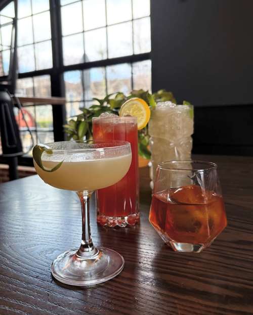 4 colorful spring cocktails in different glassware sit on a wooden table in front of a brick wall