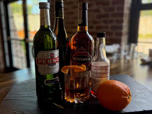 bottles pernod, rye, and brandy with a bottle of bitters behind an oldfashioned glass with a cocktail and garnish