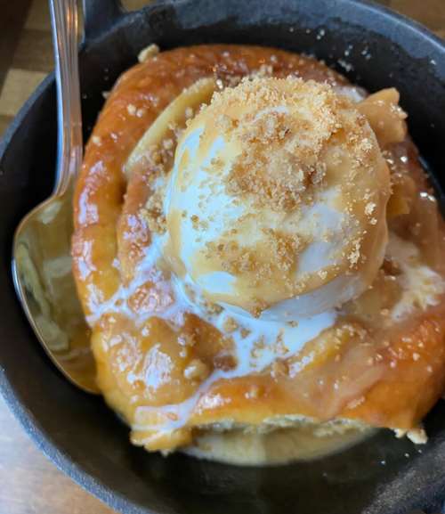 closeup of a cinnamon bun topped with vanilla icecream with serving spoon in a cast iron pan