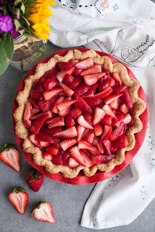a strawberry pie sits in a red pie dish with a white embroidered napkin to it's right 